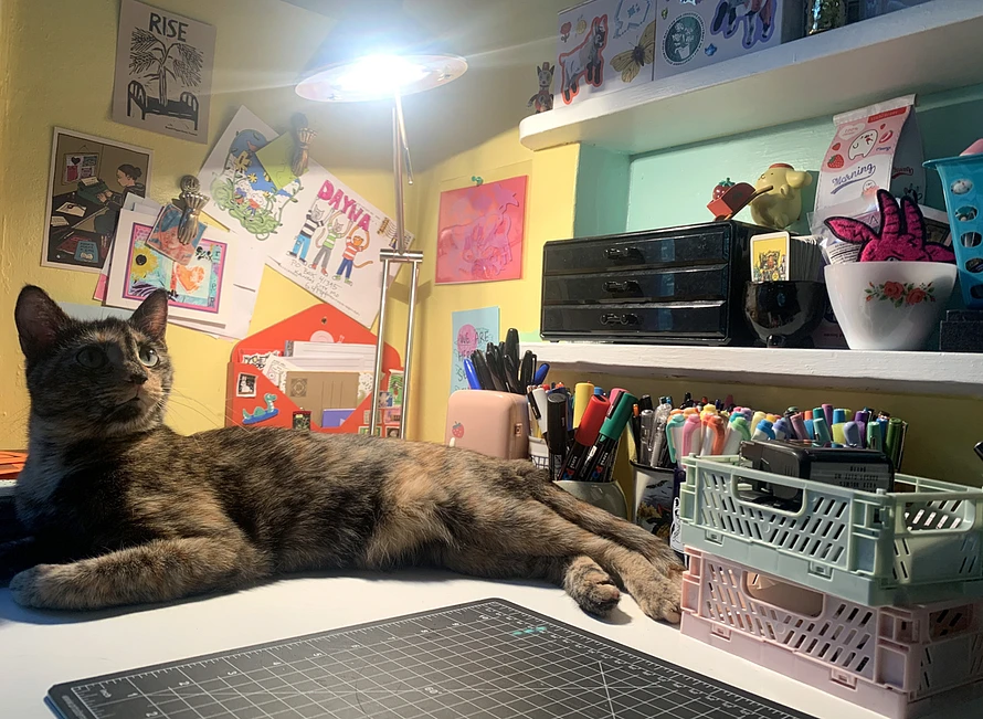 A tortie cat sits on a desk cluttered with pens and supplies. There is a bright desk lamp above the cat, partially illuminating her. In the foreground is the edge of a black cutting mat.