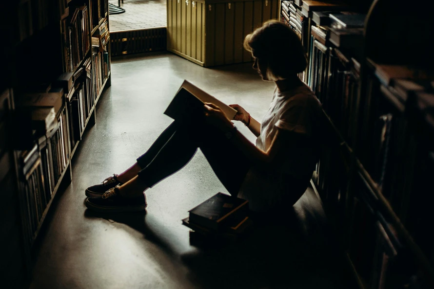 فتاة جالسة على الأرض في مكتبة هادئة، تقرأ كتابًا بينما تحيط بها رفوف مليئة بالكتب. تُظهر الصورة أجواءً دافئة ومريحة، مع إضاءة خافتة تعكس أجواء العزلة والتركيز المثالي لمحبي القراءة