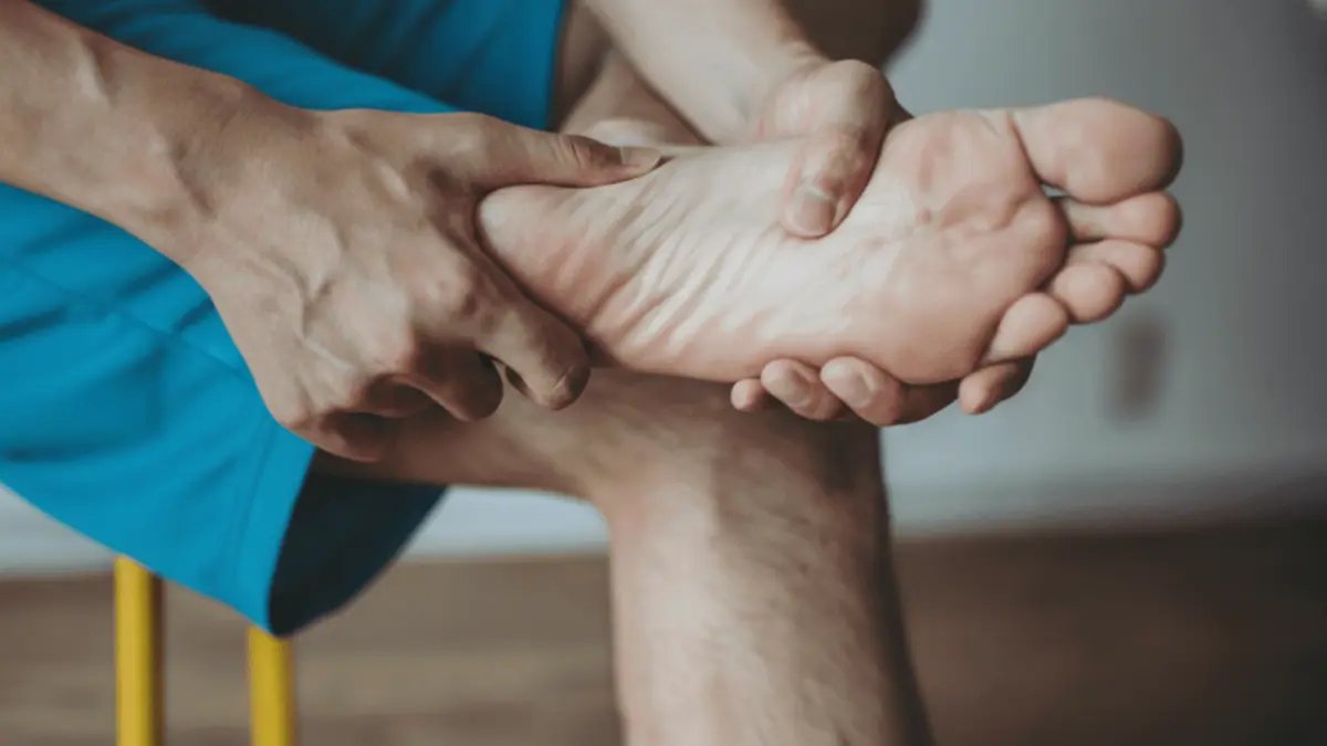Man experiencing sudden foot pain, specifically heel pain and potential plantar fasciitis, holding and massaging his foot while sitting on a chair. This image is suitable for articles and resources discussing foot pain, plantar fasciitis symptoms, and treatment options.