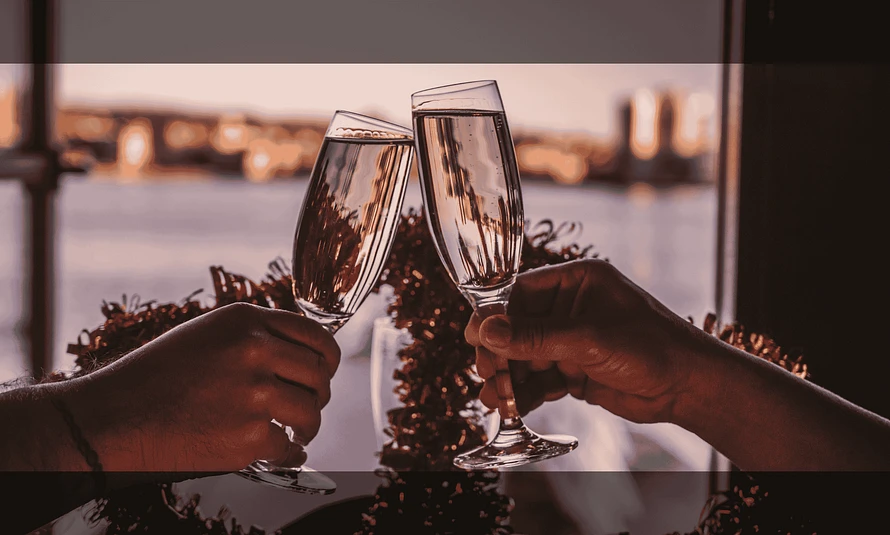 Two champagne flutes, filled with a light-colored beverage, are positioned in the foreground. They appear to be clinking or about to clink. The stems and bases of the glasses are visible.  A garland of reddish-brown tinsel or similar material sits behind the glasses, partially obscuring a third, out-of-focus glass.  In the background, a blurred cityscape at dusk or dawn is visible, with buildings silhouetted against a soft, reddish-pink sky. The light suggests a sunset or sunrise. The overall color palette is warm, with reds, golds, and soft pinks dominating. The scene suggests a celebratory or romantic occasion.