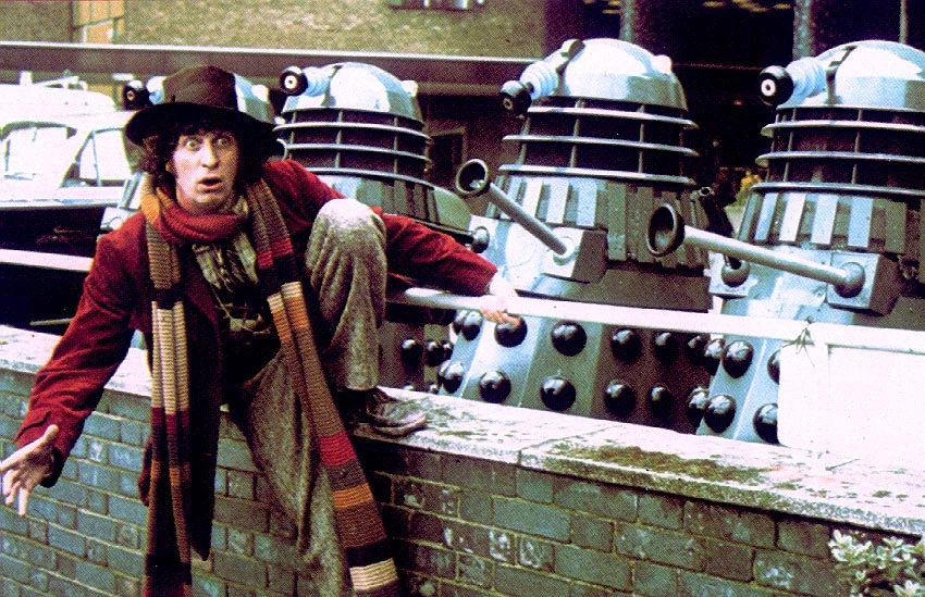 The Fourth Doctor looks shocked and hangs from a wall as Daleks approach