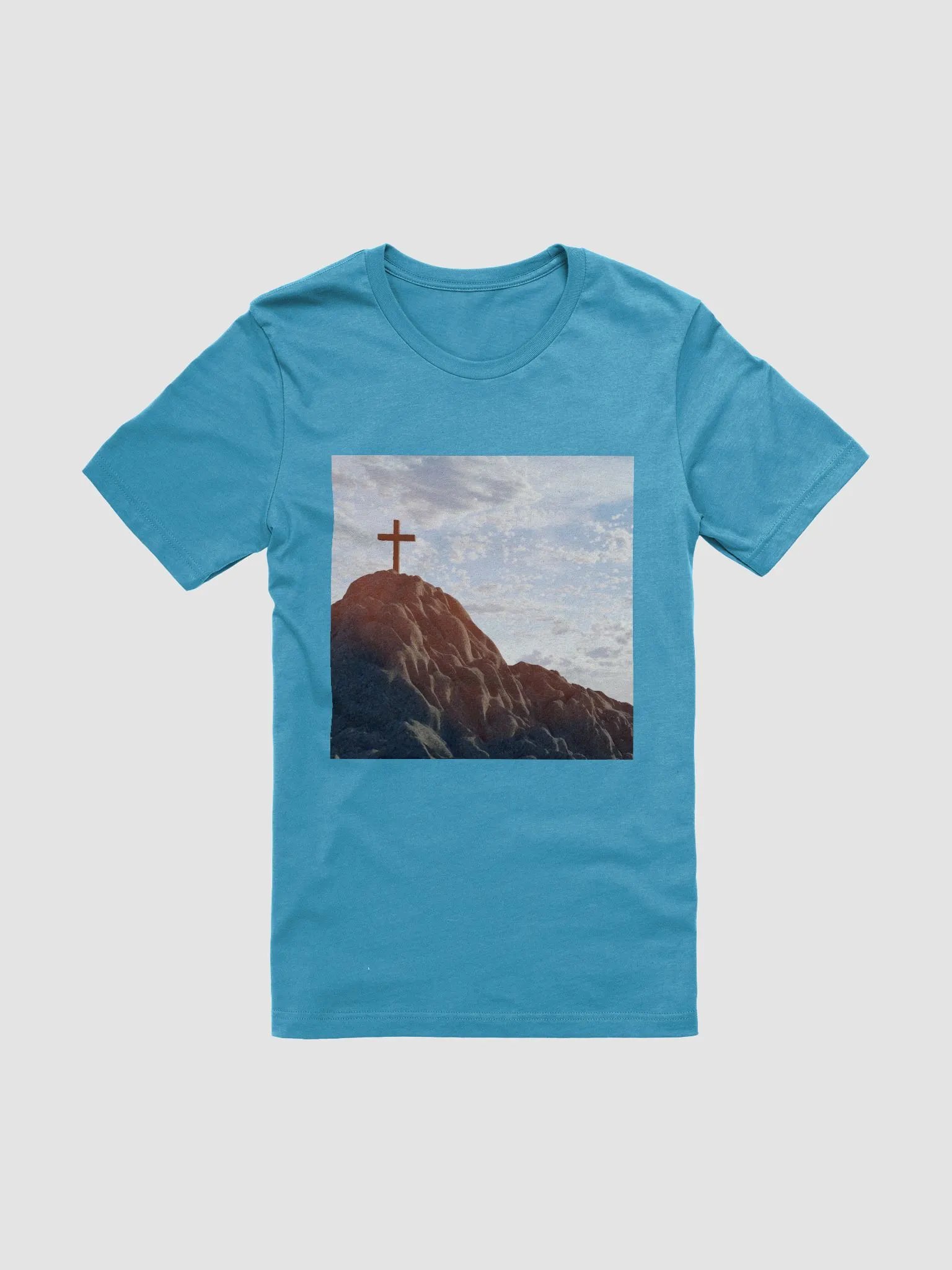 Wooden cross standing atop a rocky hill or mountain, under a sky filled with soft clouds and warm sunlight t-shirt product image (1)